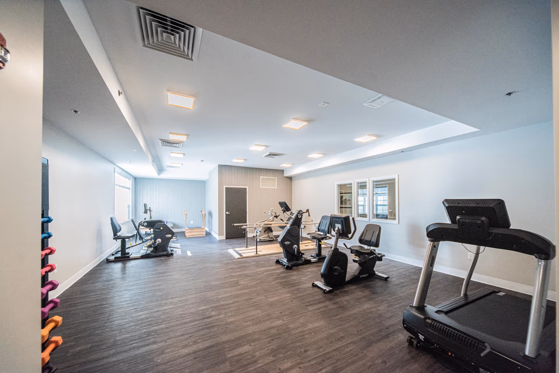 Bright fitness room with treadmills, recumbent bikes, other exercise machines and a rack of colorful dumbbells.