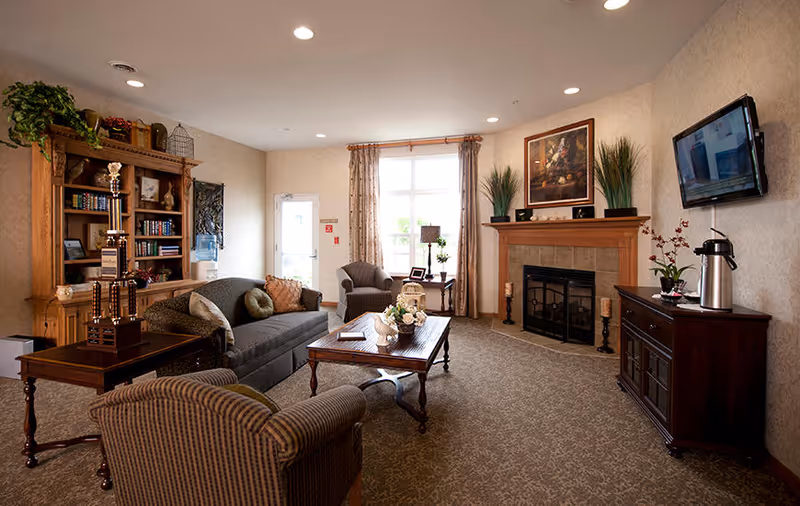 A cozy living room in Countryside Meadows featuring a gray sofa with pillows, two armchairs, a wooden coffee table with a floral centerpiece, a wooden bookshelf filled with books and decorative items, a fireplace with a framed painting above it, a flat-screen TV mounted on the wall, and a wooden cabinet with a coffee dispenser and a plant. The room has beige walls, carpeted floor, and a large window with curtains letting in natural light.