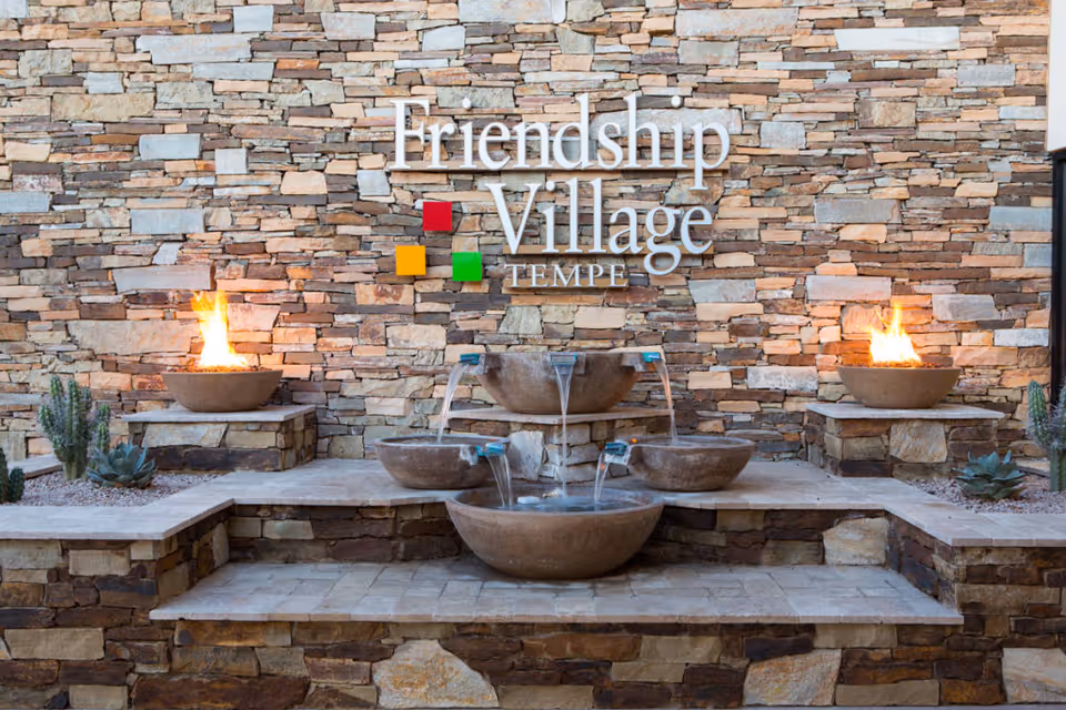 Stone wall with the Friendship Village Tempe logo mounted on it, featuring colorful square accents. In front of the wall is a tiered stone water fountain with water flowing from the top bowl to the lower bowls. On either side of the fountain are fire bowls with flames burning, surrounded by small desert plants.