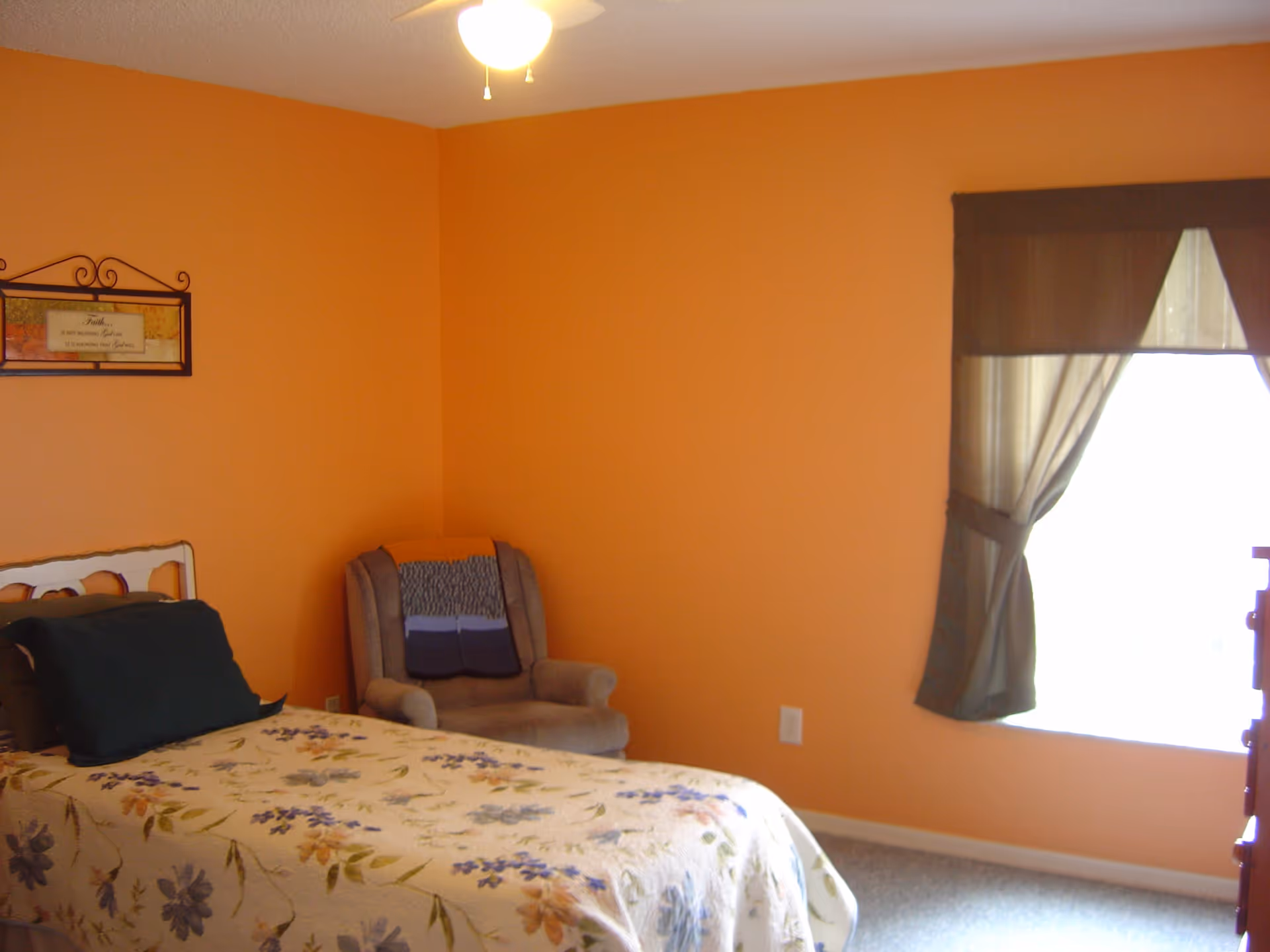 Small bedroom with orange walls, a single floral bed, an upholstered armchair, and a curtained window.