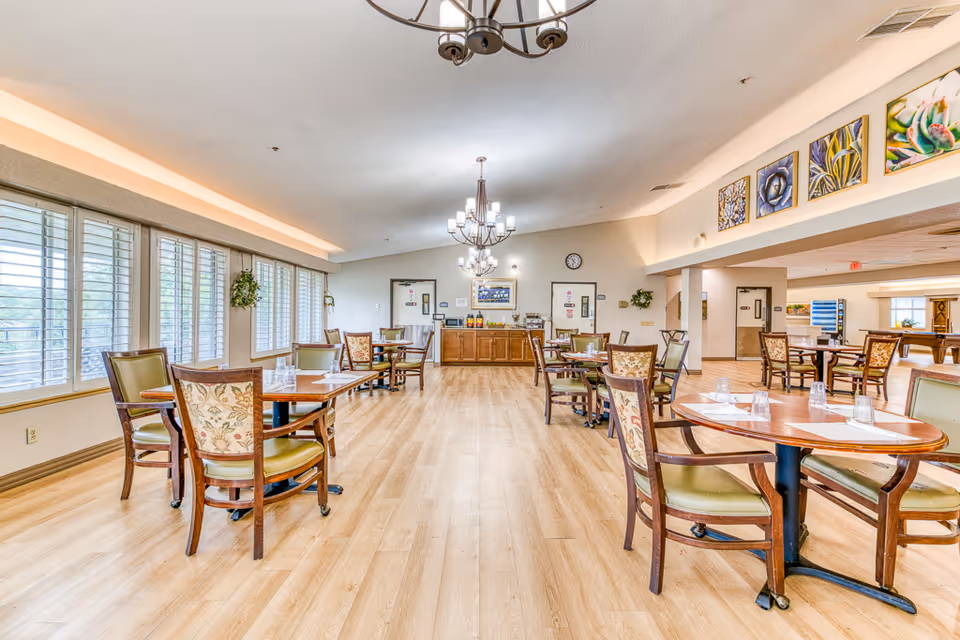 A bright and spacious dining room with wooden floors and multiple round tables surrounded by chairs. Large windows with white shutters line one side of the room, allowing natural light to fill the space. The walls are decorated with colorful paintings and plants. Two chandeliers hang from the ceiling, and a sideboard with beverages and condiments is visible at the far end of the room.