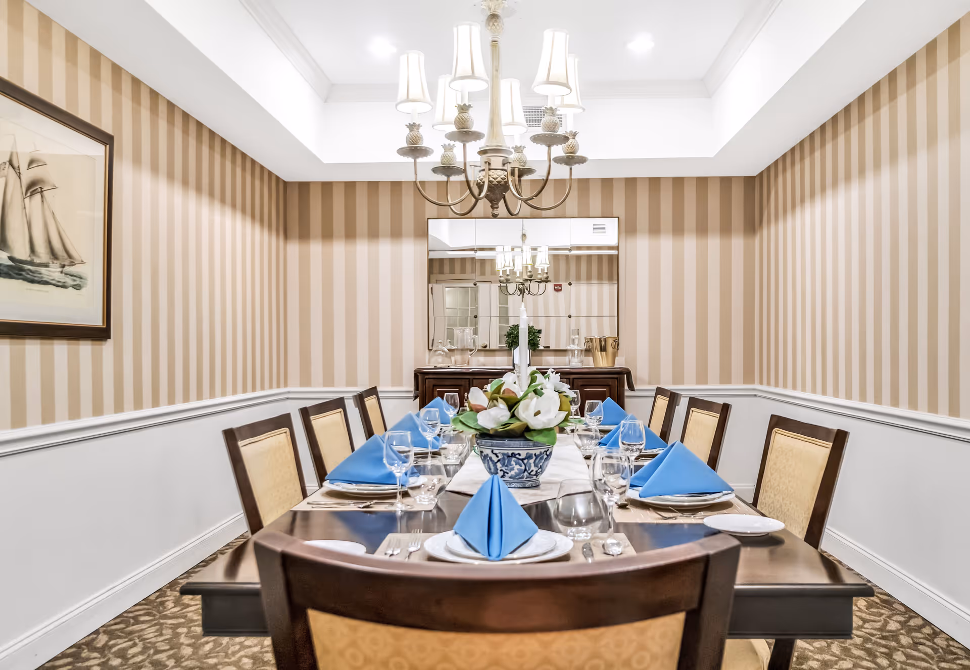 A formal dining room with a long wooden table set for eight people. Each place setting includes a white plate, silverware, a glass, and a blue folded napkin. A floral centerpiece with white flowers is in the middle of the table. The room has beige and white striped wallpaper, a chandelier with multiple lampshades hanging from the ceiling, a framed picture of a sailboat on the left wall, and a large mirror on the back wall reflecting the chandelier.