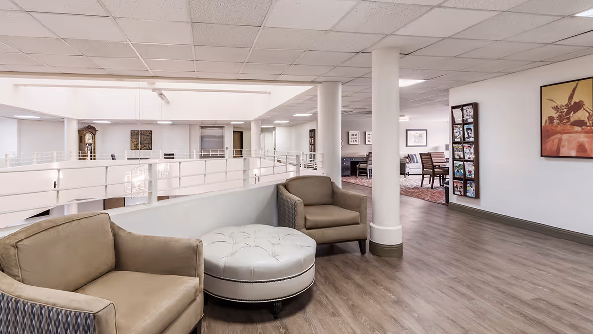 Interior view of a senior living community common area with beige armchairs and a round white ottoman on wood flooring. The space has white walls, a railing overlooking a lower level, and several columns. In the background, there is a seating area with tables and chairs, a grandfather clock, and framed artwork on the walls.