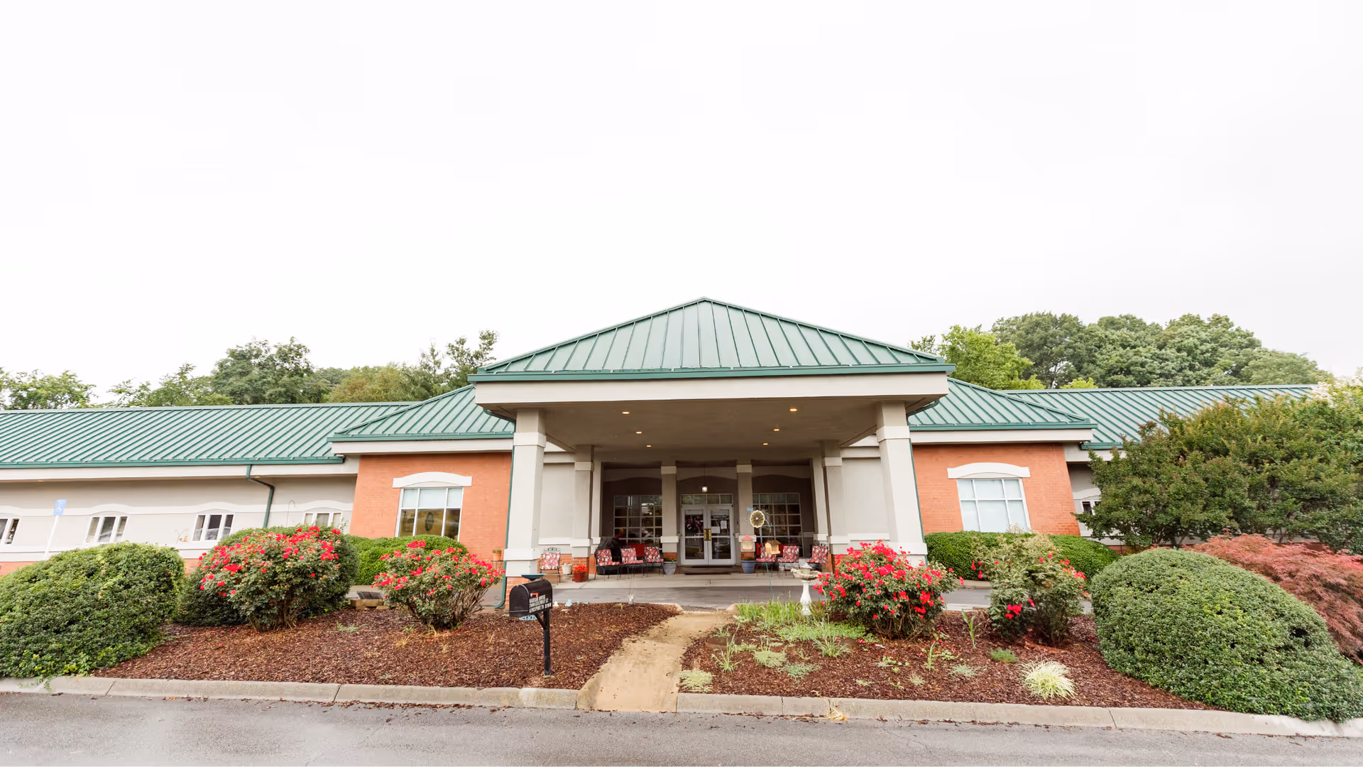 Front entrance of a single-story senior living facility with a green metal roof, covered porte-cochère, and landscaped flower beds.
