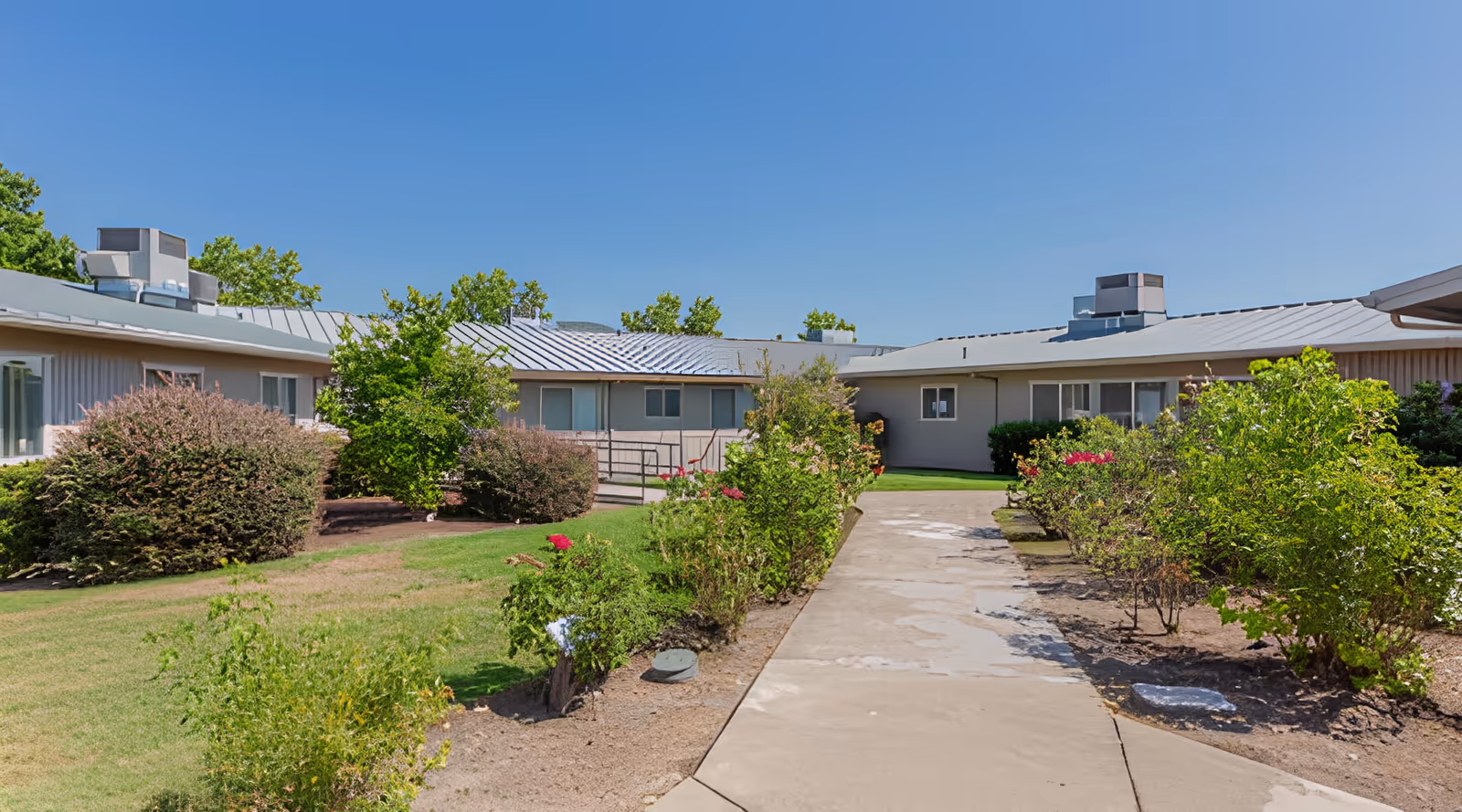Outdoor view of a single-story building with a metal roof, surrounded by green bushes and trees under a clear blue sky. A concrete pathway leads through the landscaped garden area towards the building.