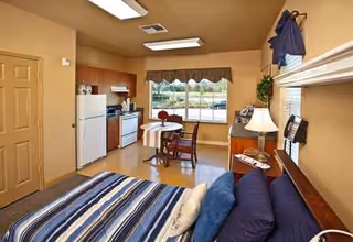 Interior of a senior living suite with bed, sofa, table, and kitchenette near window.