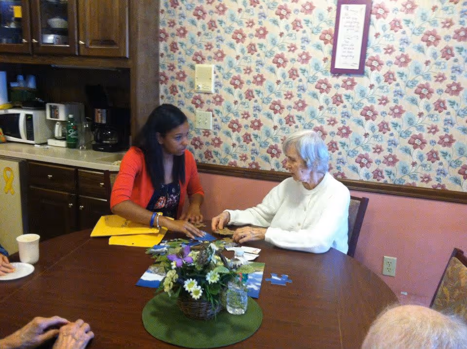 An elderly woman and a younger woman sitting at a round wooden table working on a jigsaw puzzle together in a room with floral wallpaper. The table has a green placemat with a basket of flowers and a glass of water. The background shows a kitchen area with cabinets, a microwave, and a coffee maker.