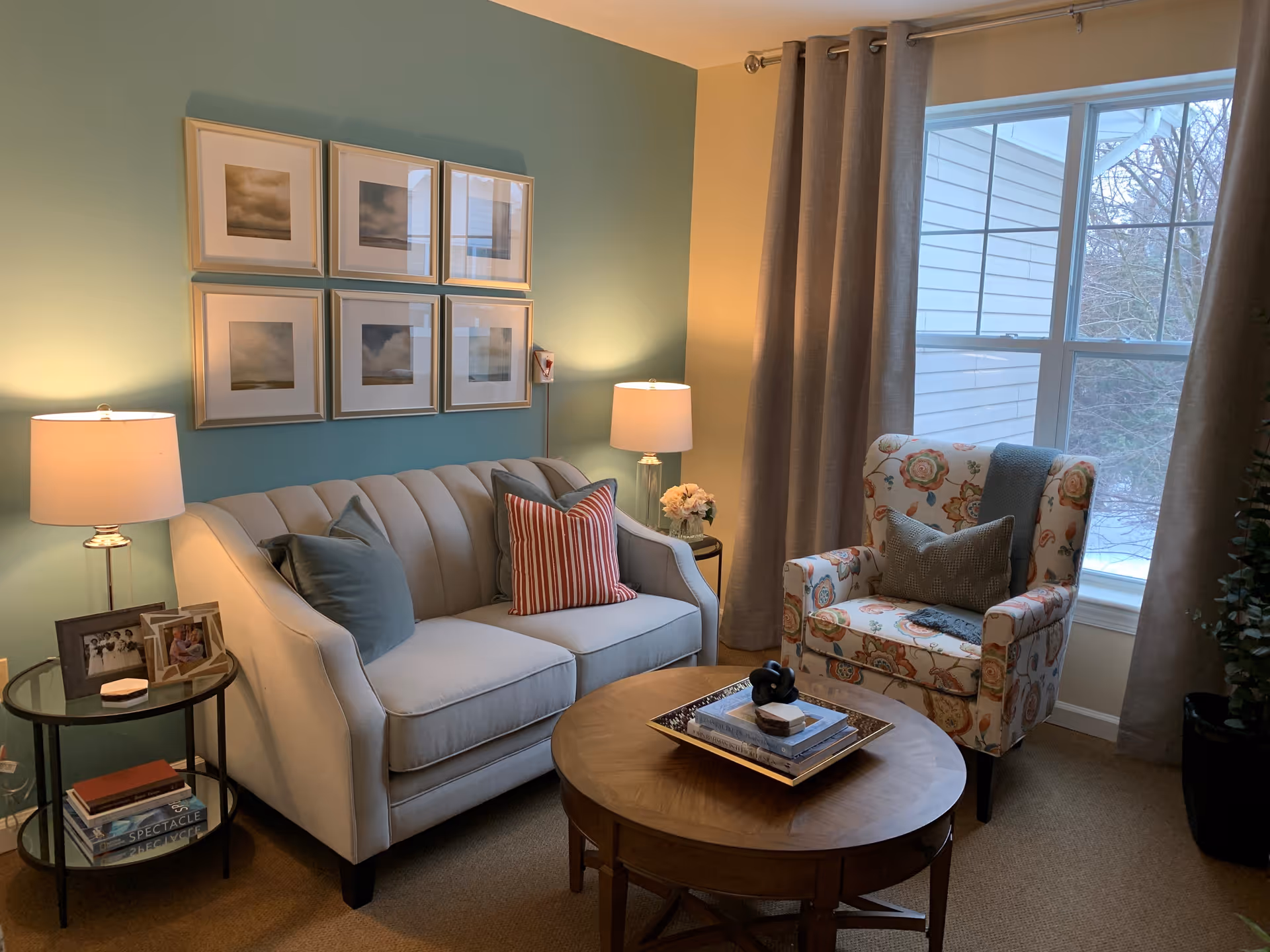 A cozy living room with a light beige sofa adorned with three pillows, a floral patterned armchair with two pillows, a round wooden coffee table with books and decorative items, two side tables each with a lamp and framed photos, and a large window with beige curtains letting in natural light. The walls are painted in a soft green and cream color, with six framed pictures hanging above the sofa.
