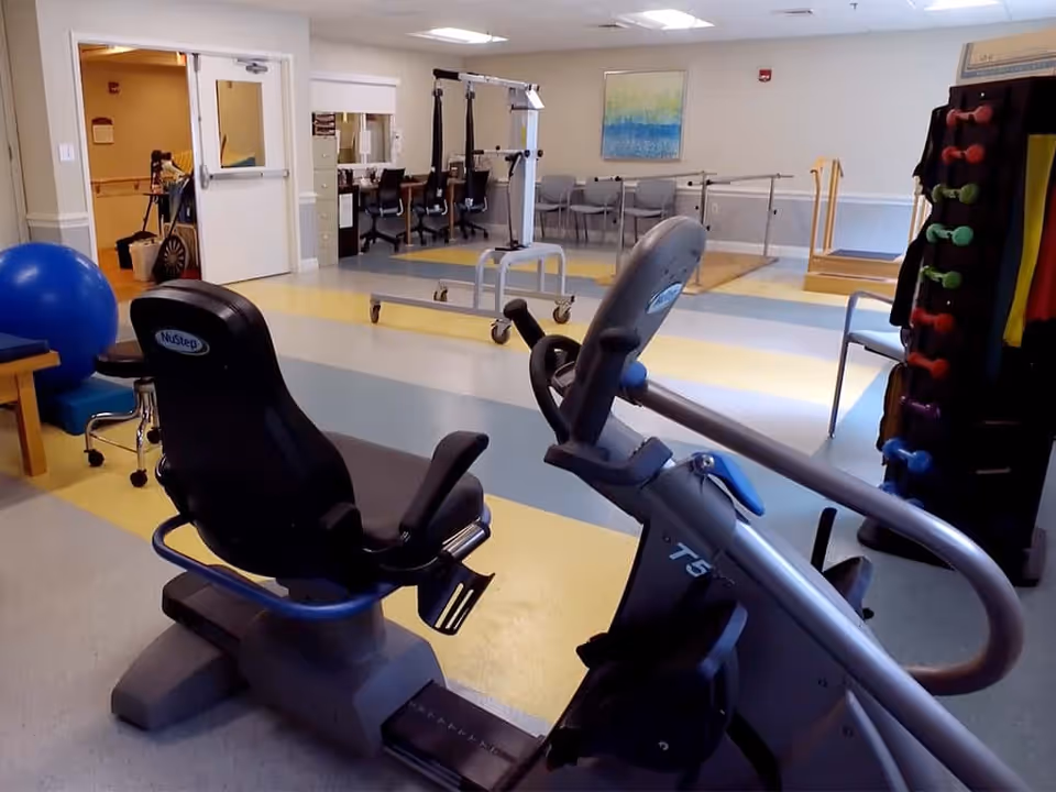 A rehabilitation or physical therapy room with exercise equipment including a NuStep recumbent bike, parallel bars for walking practice, a rack of colorful dumbbells, chairs along the wall, and a large blue exercise ball. The room has a clean, spacious layout with light-colored walls and a painting on the far wall.