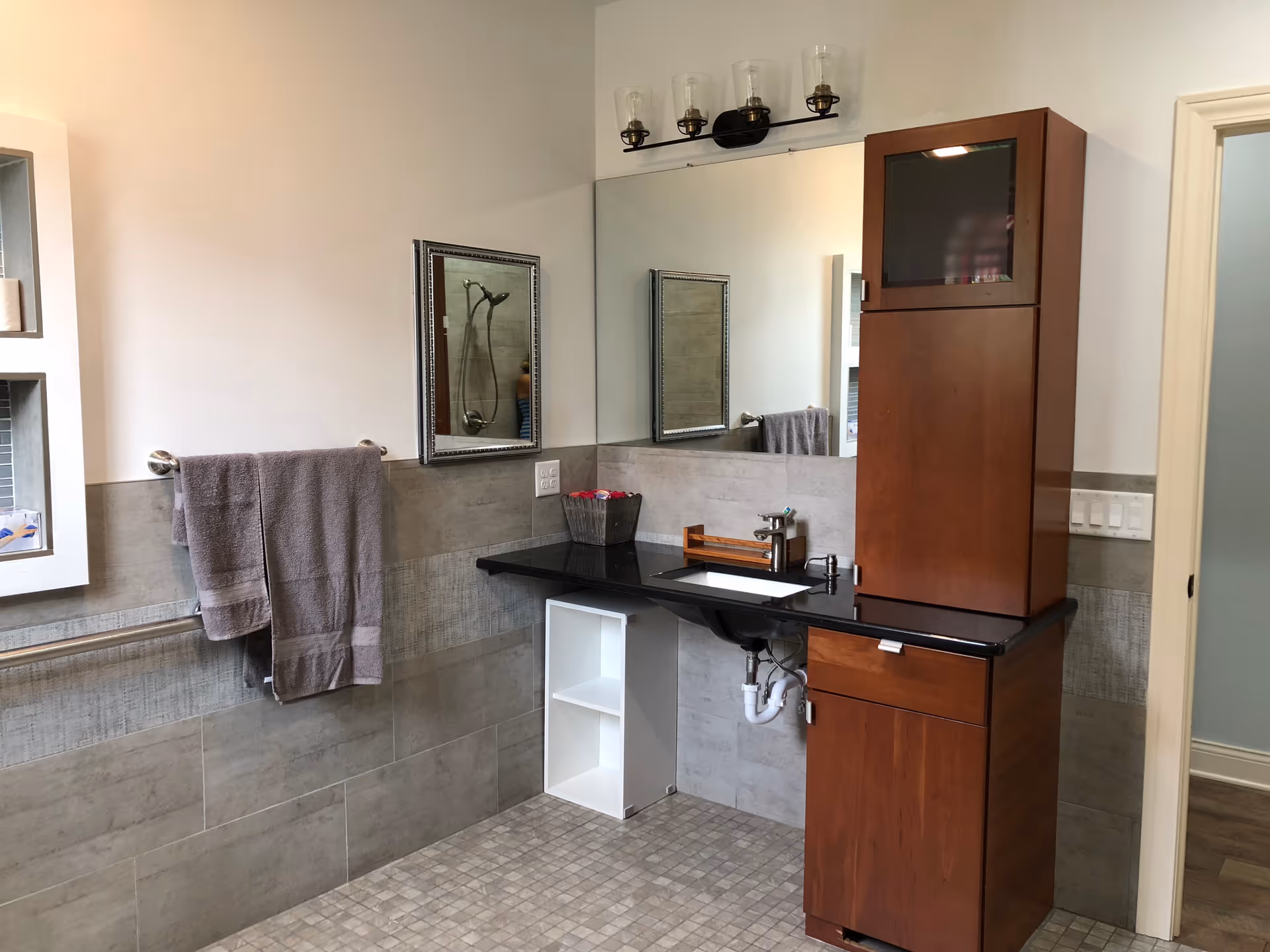 A modern bathroom with a black countertop sink, tall wooden storage cabinet, large mirror, gray towels on a rail, and tiled floor and walls.
