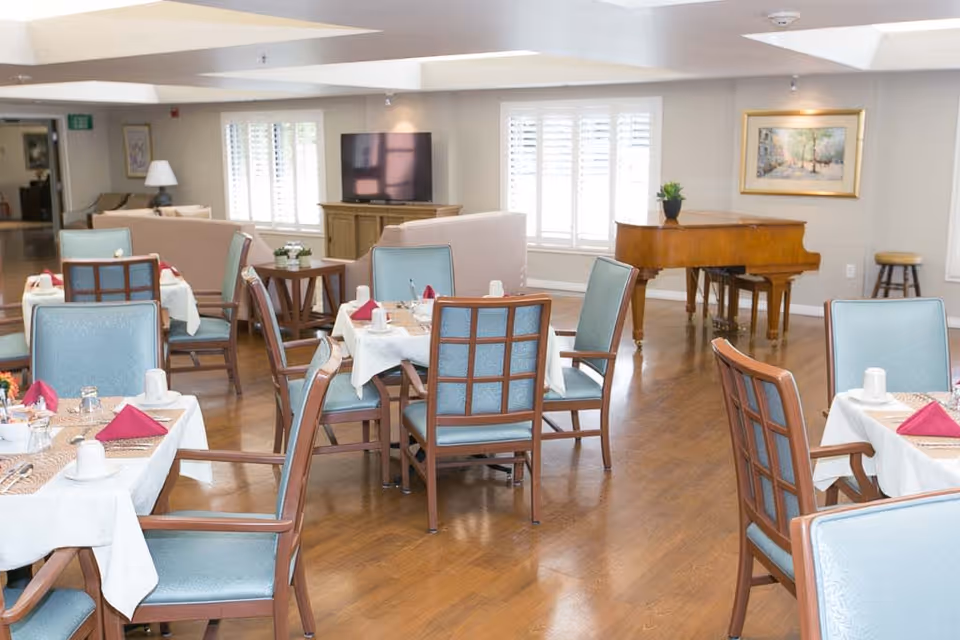A bright dining room in a senior living facility with multiple tables set with white tablecloths, red napkins, cups, and silverware. The room features light blue cushioned chairs, wooden flooring, a grand piano in the corner, a television on a wooden stand, and large windows with white shutters allowing natural light to fill the space.
