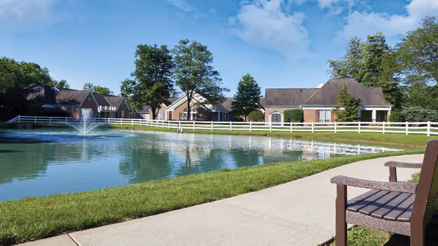 A paved walkway and bench beside a pond with a fountain, white fence, and brick residential buildings under a blue sky.