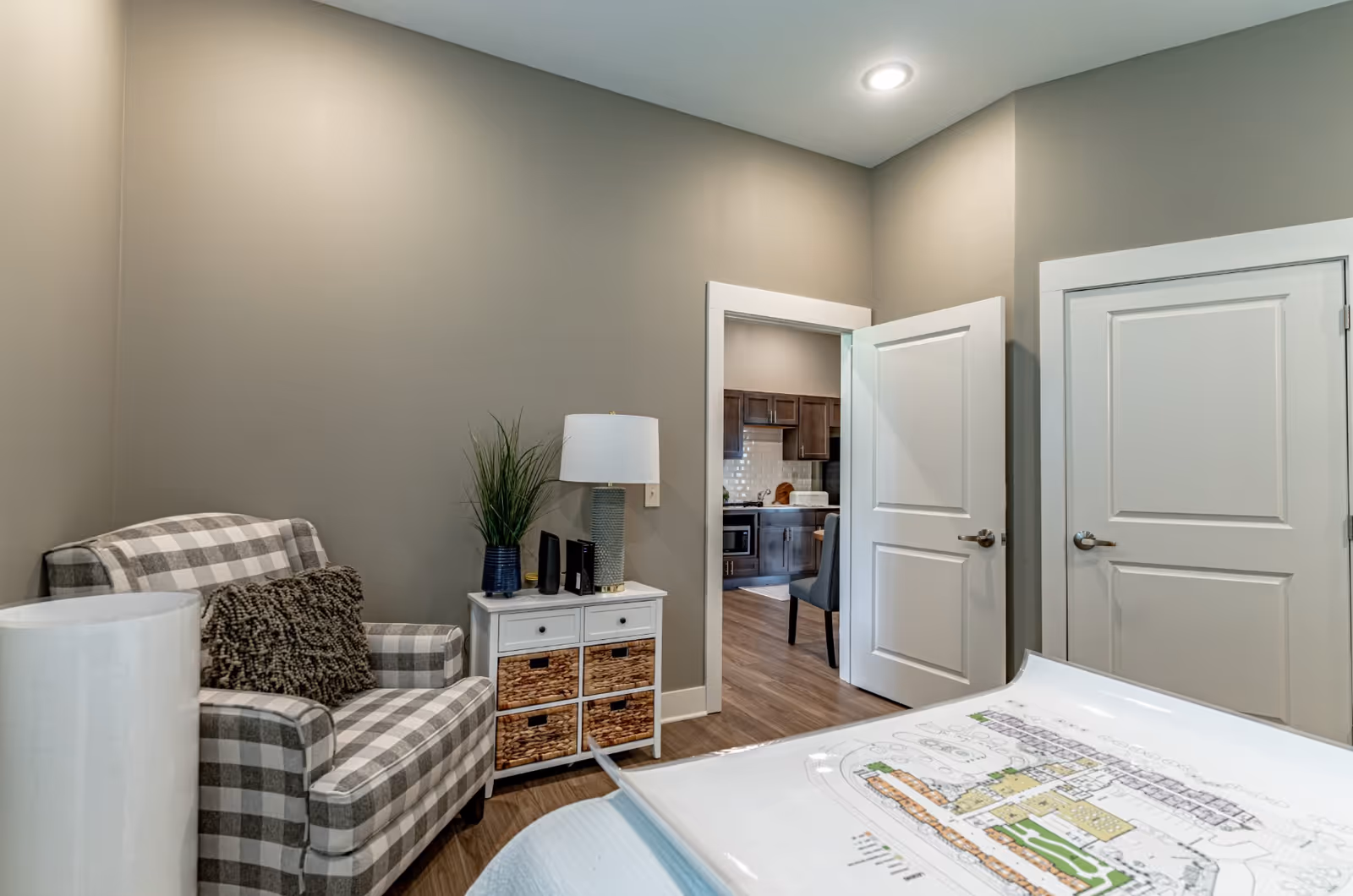 Cozy interior room with a plaid armchair, a white side table with lamp and baskets, and open doors revealing a kitchen.