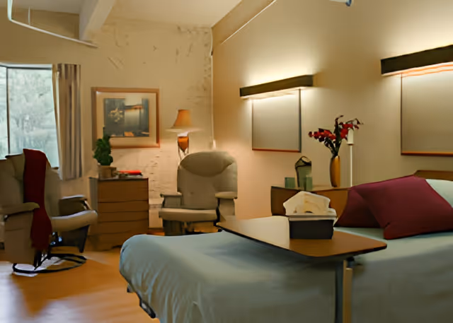A cozy senior facility bedroom with a bed and over-bed table, upholstered armchairs, a dresser, lamp, and soft wall lighting.