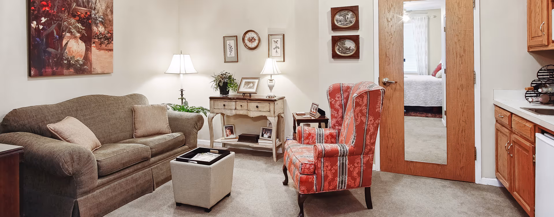 Cozy living room with a sofa, patterned armchair, ottoman, side tables with lamps, artwork, and a doorway leading to a bedroom.