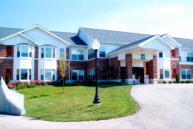 Exterior view of a two-story retirement community building with white and red brick facade, large windows, a covered entrance with rocking chairs, a street lamp, and a well-maintained lawn with small trees and bushes.