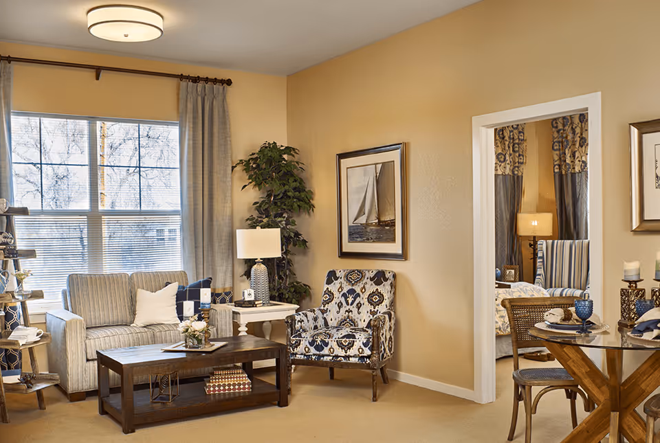 Bright, warmly decorated living room with a sofa, patterned armchair, coffee table, side tables and a view into an adjacent room.