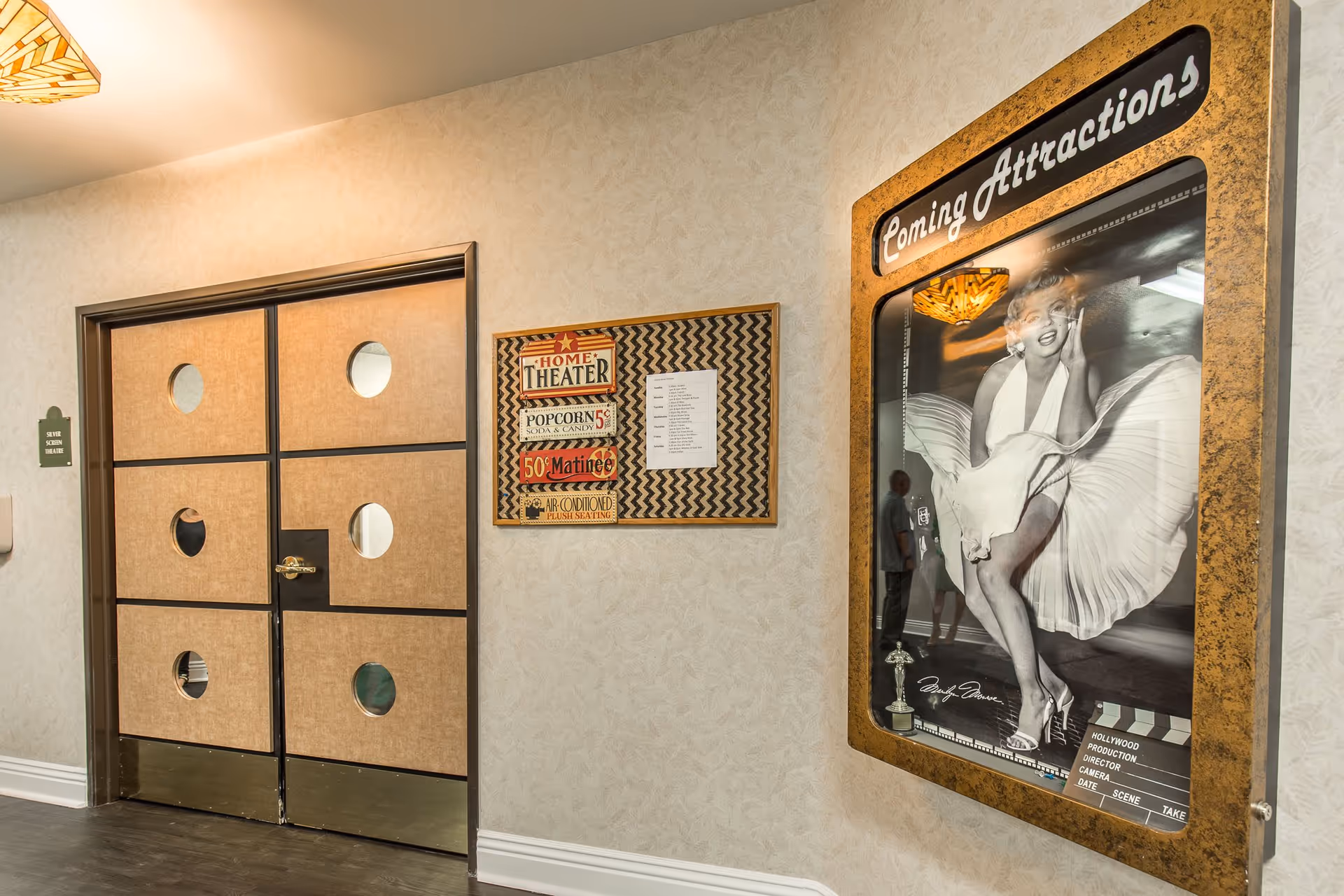 Interior hallway of Windsor Court Assisted Living featuring double doors with circular windows labeled 'Seven Screen Theatre.' Next to the doors is a bulletin board with a vintage-style sign advertising home theater popcorn, soda, and candy, and a schedule. On the right wall is a framed black and white poster of Marilyn Monroe with the heading 'Coming Attractions.'