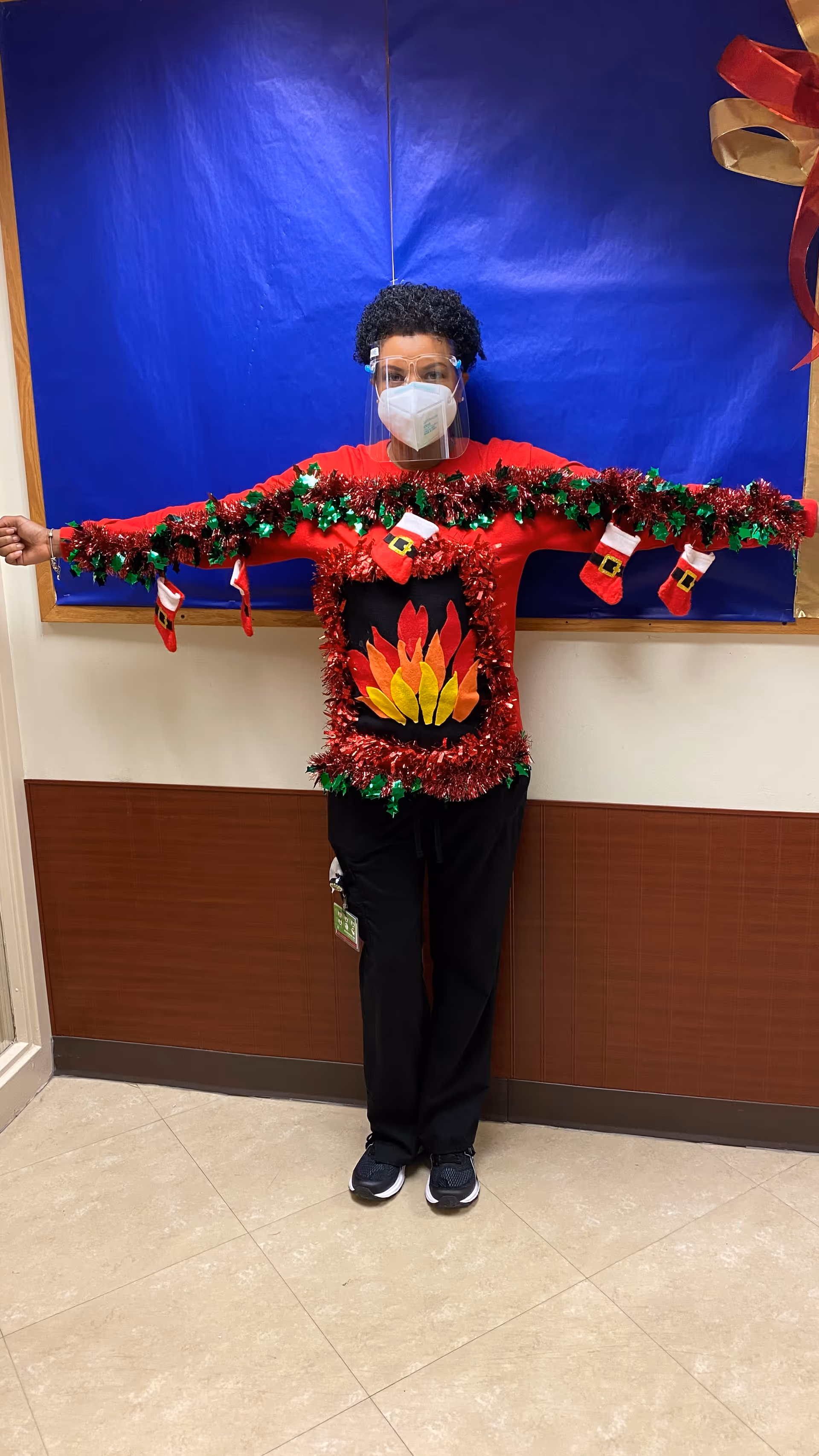 A person wearing a festive red sweater decorated with tinsel, small Christmas stockings, and a flame design on the front. The person is also wearing a face mask and a clear face shield, standing with arms outstretched in a hallway with a blue bulletin board behind them.