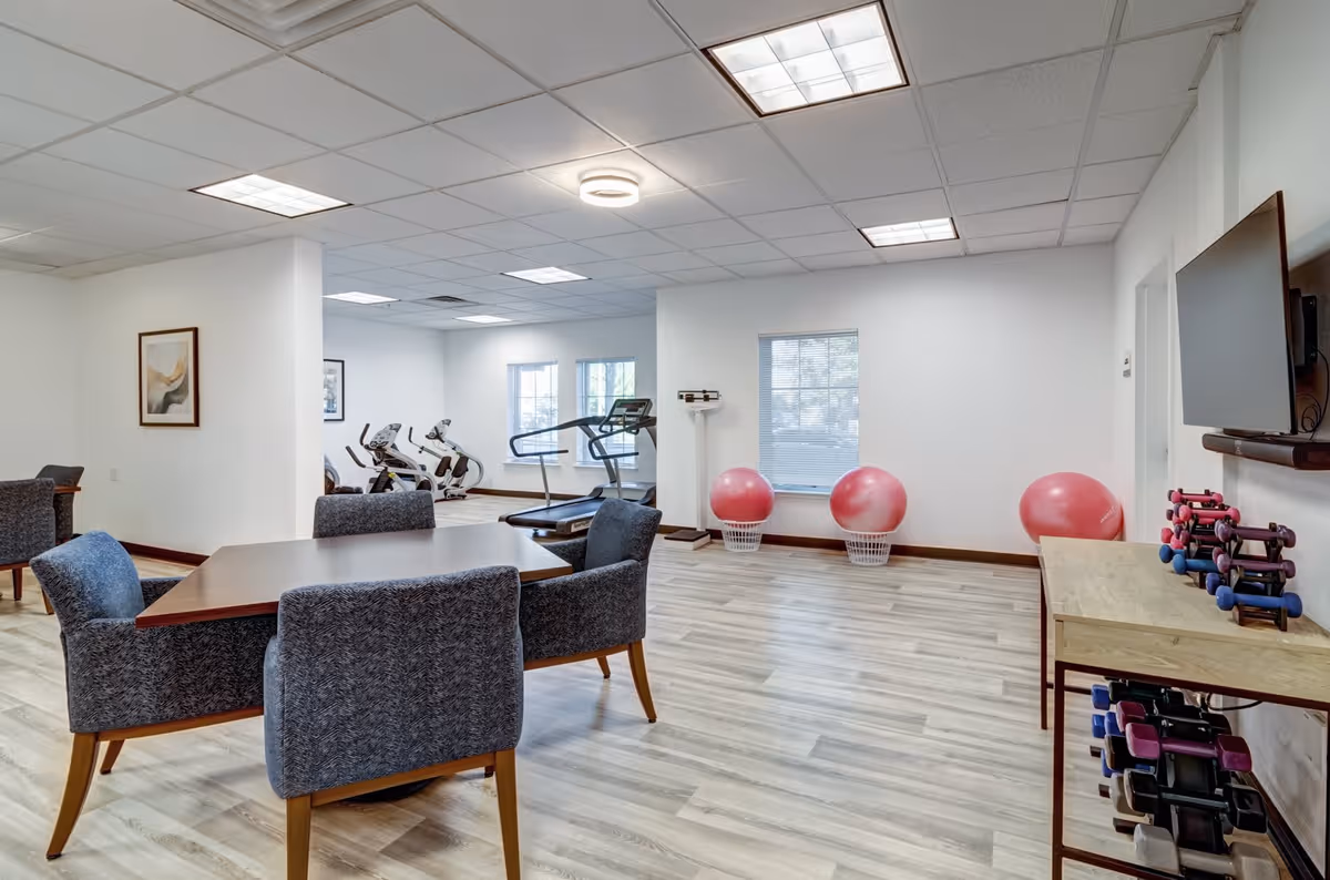 A bright room with light wood flooring featuring a table with four upholstered chairs in the foreground. In the background, there are exercise machines including a treadmill and stationary bikes, along with pink exercise balls and a scale near a window. A TV is mounted on the wall above a rack holding various dumbbells.