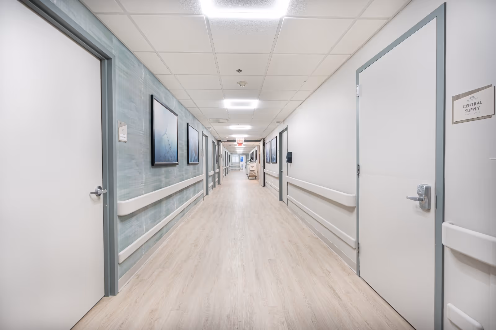 Long, well-lit hospital-style hallway with handrails, closed doors, and framed artwork along the walls.