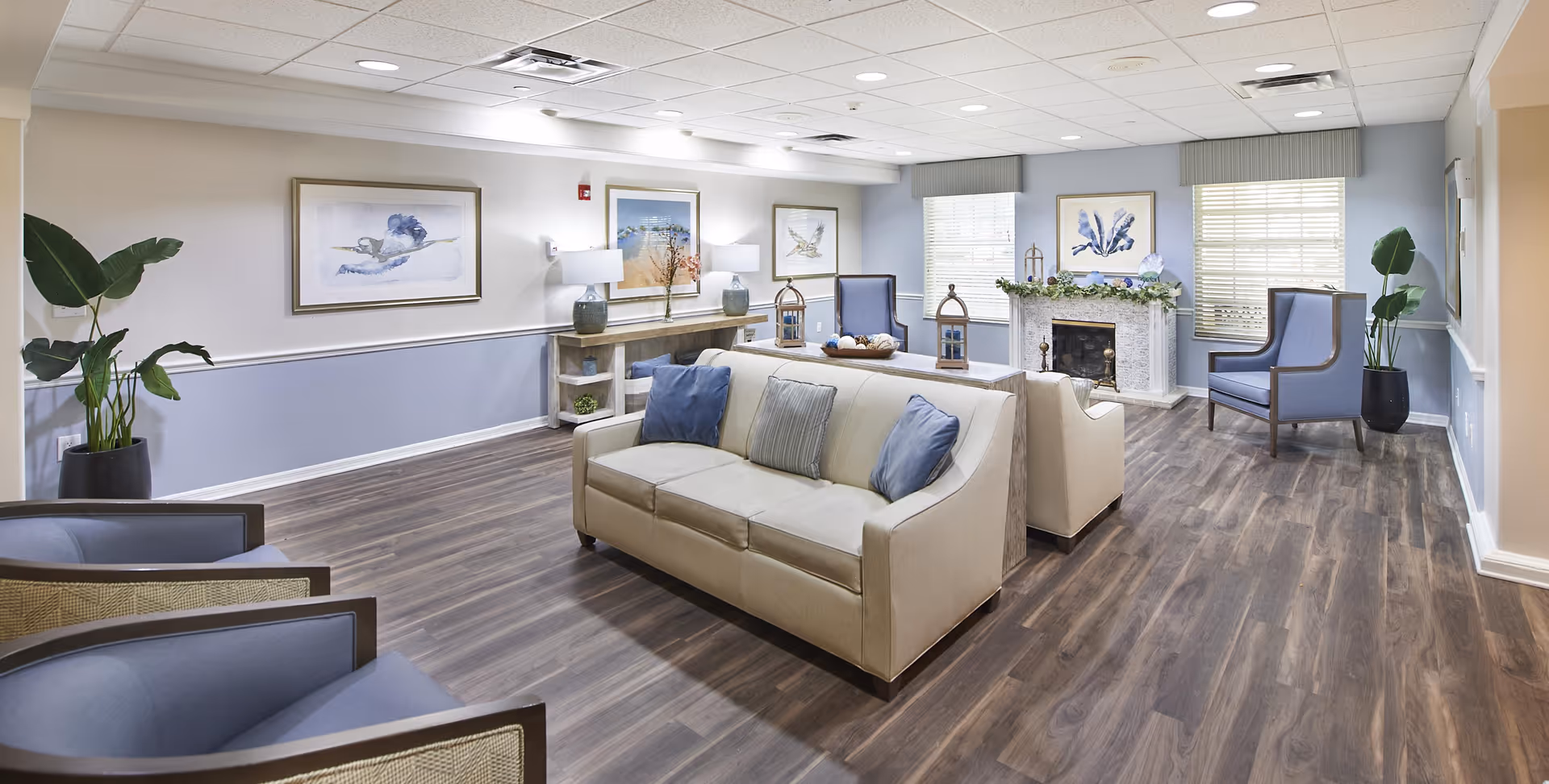 Communal living room with sofas and armchairs arranged around a fireplace, wood-look flooring, artwork, and potted plants.