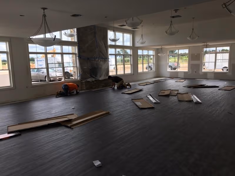 A large room with multiple windows letting in natural light, two workers kneeling on the floor installing dark wood-like flooring, several pieces of cardboard and flooring materials scattered around, and a covered fireplace in the background.