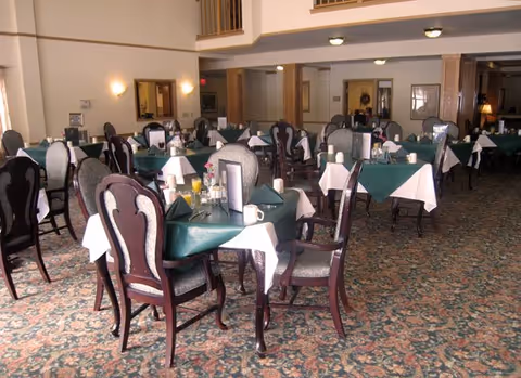 A spacious dining room with multiple tables covered with white and green tablecloths, set with glasses, cups, and napkins. The room has patterned carpet flooring, wooden chairs with cushioned seats, and soft lighting from wall sconces and ceiling lights.
