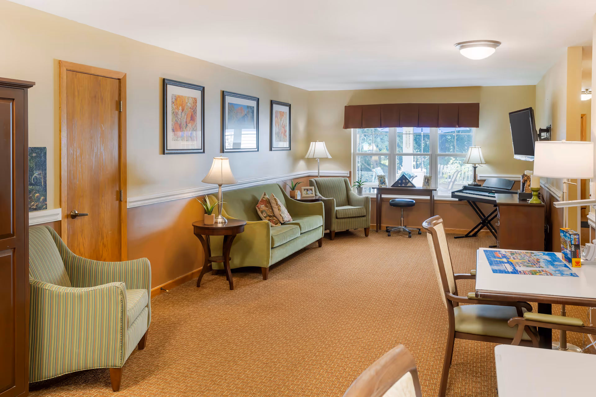 A cozy living room area in Brookdale Battle Creek featuring a green sofa with patterned pillows, a matching armchair, and a wooden side table with a lamp. The room has beige walls with three framed artworks, a window with a brown valance, a desk with a swivel chair, a keyboard on a stand, and a wall-mounted TV. There are additional chairs and tables in the foreground, and the room is well-lit with ceiling and table lamps.