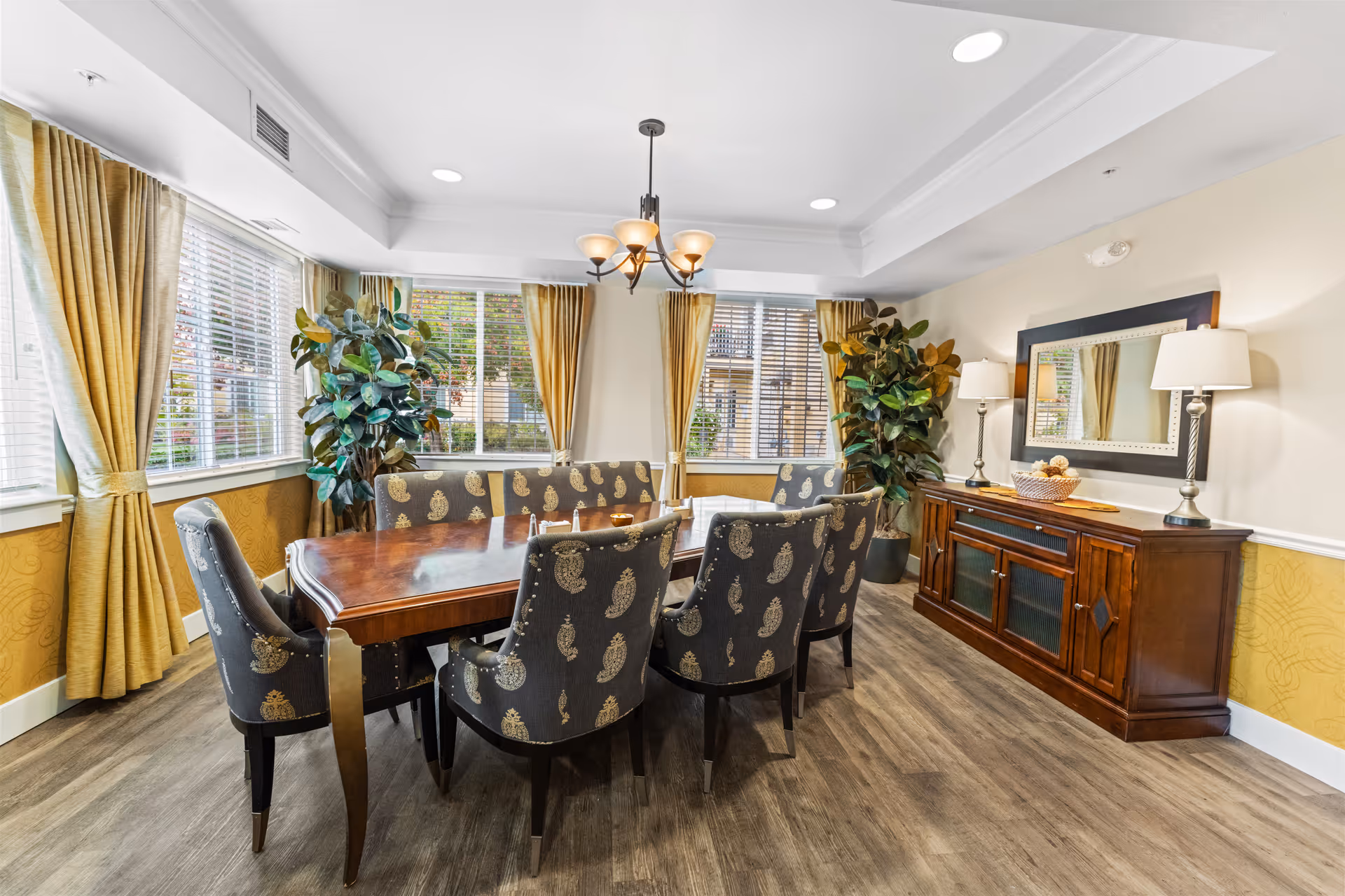 A well-lit dining room with a wooden dining table surrounded by six upholstered chairs with a paisley pattern. The room features large windows with yellow curtains, two large potted plants in the corners, a wooden sideboard with two lamps and a decorative basket, and a large mirror above the sideboard. The floor is wood, and a chandelier hangs from the ceiling.