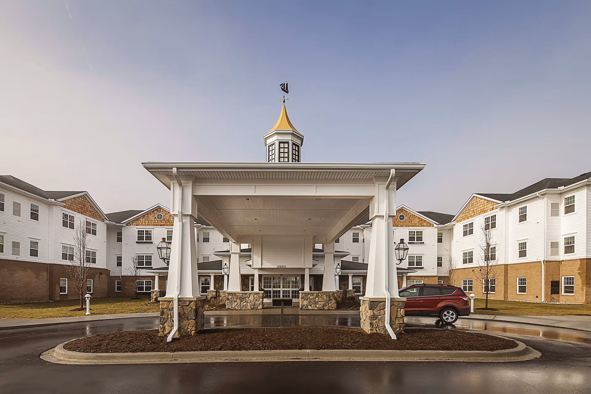 Front exterior view of American House Lakeshore facility featuring a covered entrance with white pillars and stone bases, a cupola with a weather vane on top, and a red vehicle parked on the right side. The building has white siding with brown shingle accents and multiple windows.