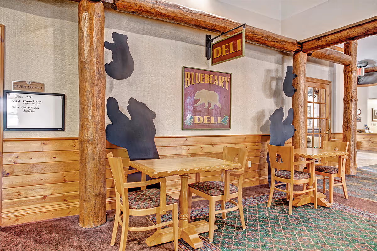 Interior view of a rustic deli area named Bluebeary Deli with wooden tables and chairs. The walls are decorated with bear-shaped cutouts and a sign featuring a bear and the deli name. The space has wooden beams and paneling, with a carpeted floor and a whiteboard menu on the wall.