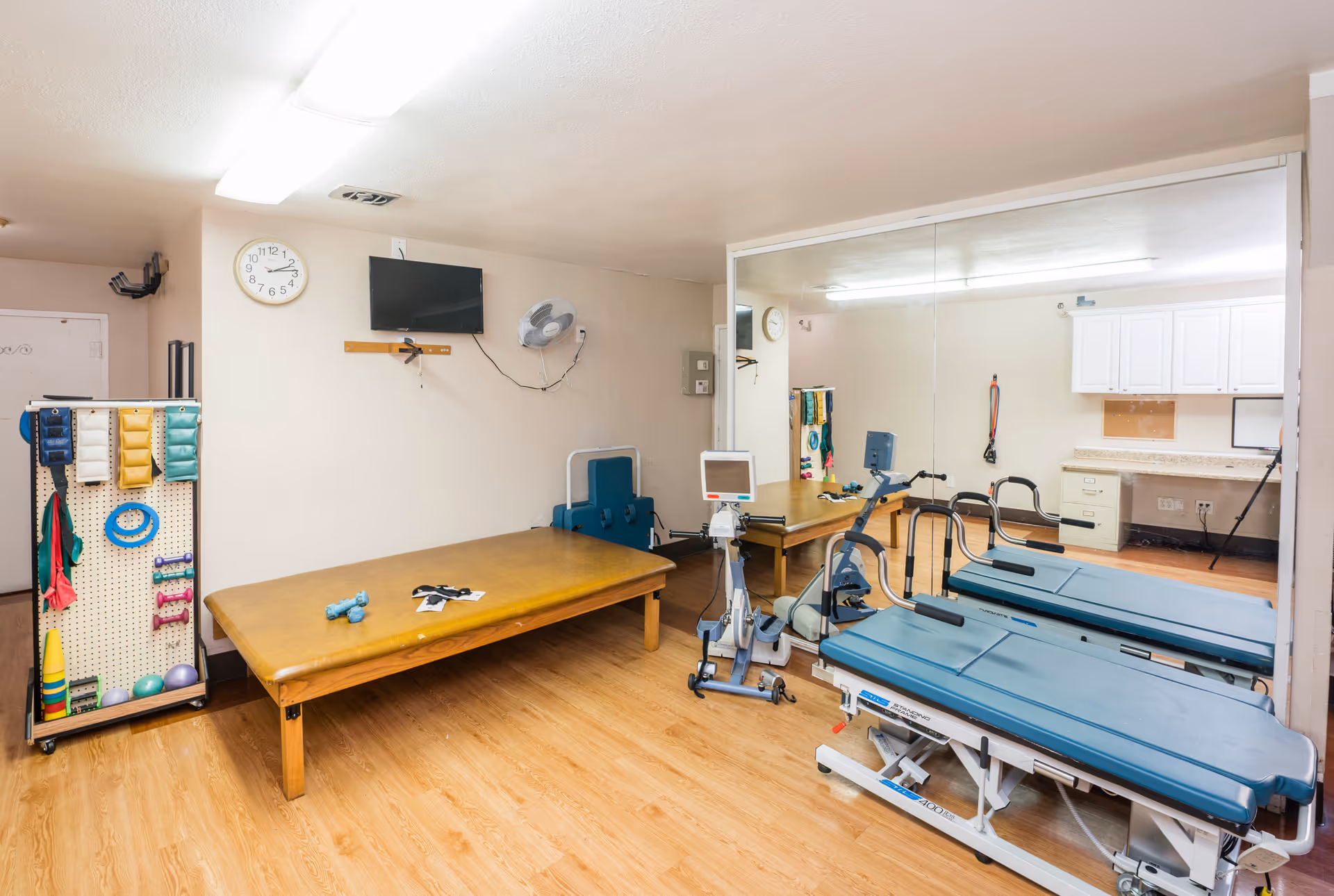 A physical therapy room with a padded treatment table, exercise equipment including a stationary bike and a treadmill, a rack with various exercise bands and small weights, a wall-mounted TV, a clock, and a large mirror covering one wall. The room has wooden flooring and white walls with overhead fluorescent lighting.