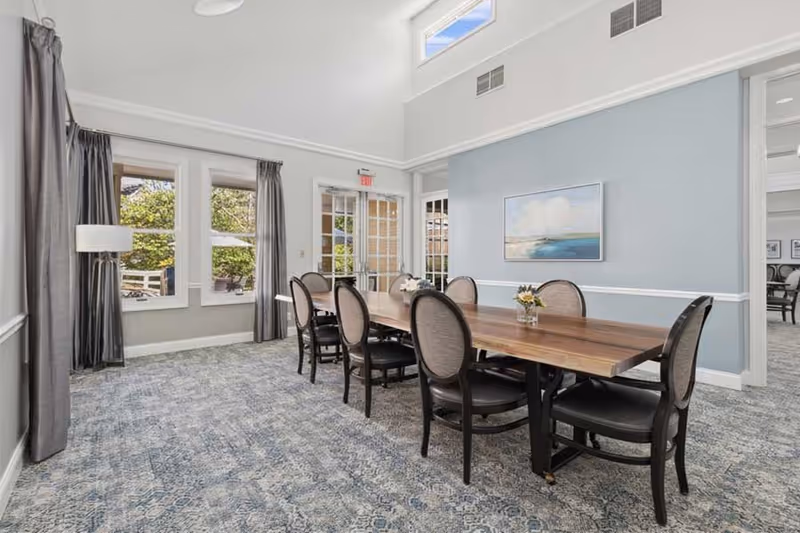 A bright dining room with a long wooden table surrounded by eight upholstered chairs. The room has large windows with gray curtains, a light blue wall with a landscape painting, and a patterned carpet. There is a door leading outside and another room visible through an open doorway.