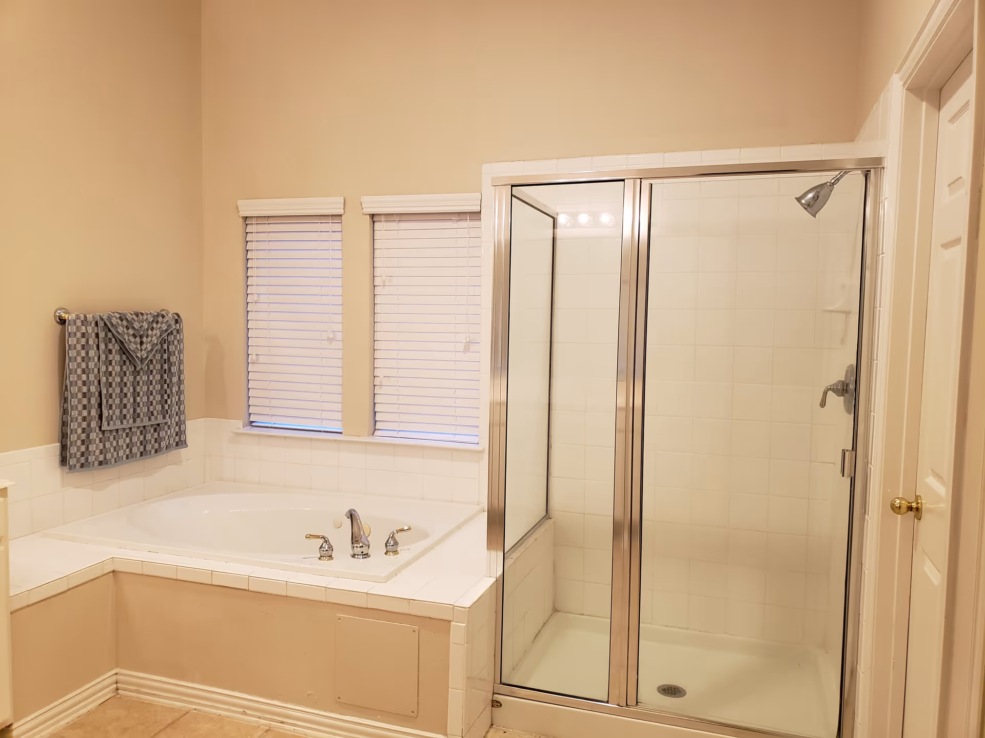 Bathroom with a corner soaking tub, two small windows with blinds, a towel on a wall rack, and a glass-enclosed shower.