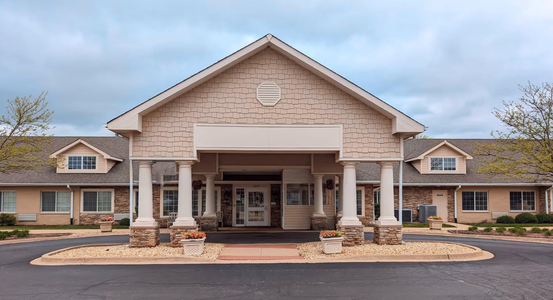 Front exterior view of Rock Run Memory Care facility showing a covered entrance with columns, flower pots, and a paved driveway under a cloudy sky.