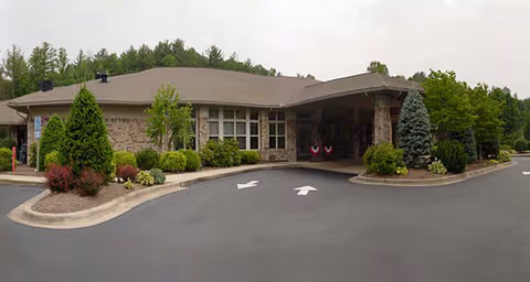 Exterior view of a single-story brick building with a covered entrance and landscaped surroundings including bushes and trees. The driveway in front has directional arrows painted on the asphalt.