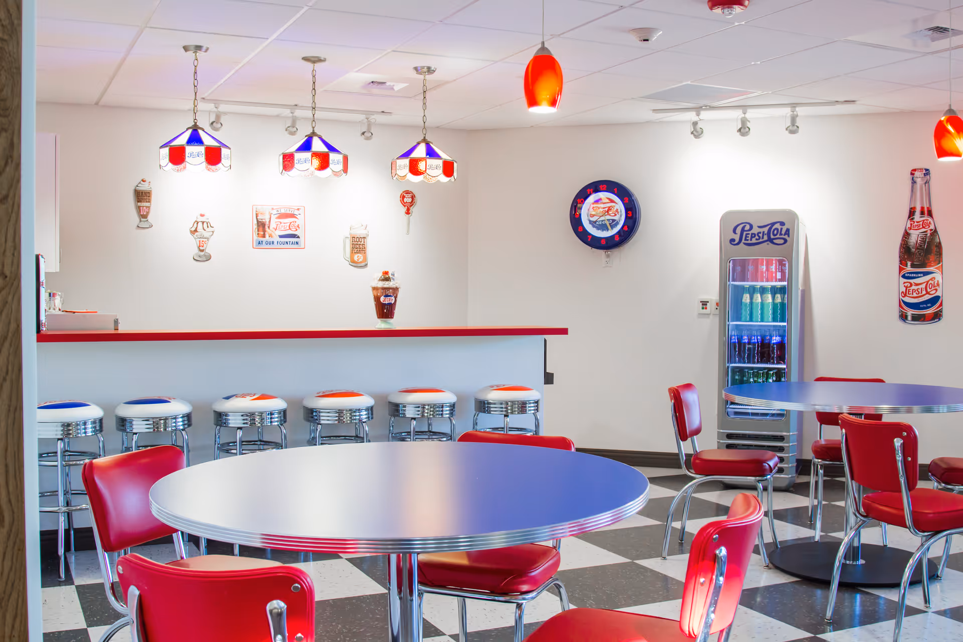 Retro-themed dining area with red chairs, round tables, a counter with barstools, and a Pepsi cooler.