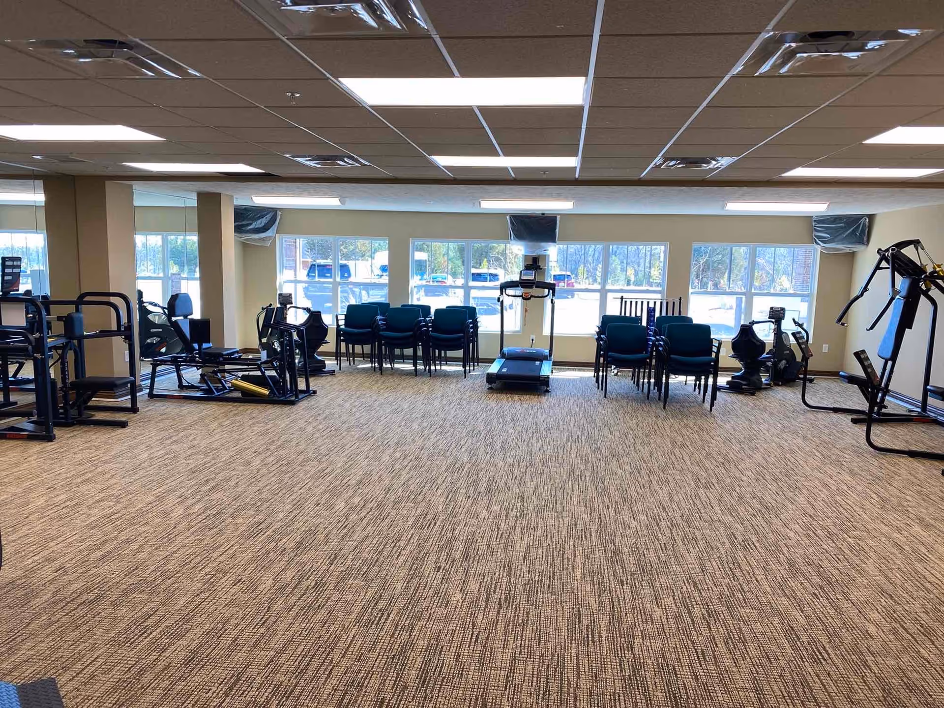 A spacious fitness room with various exercise equipment including a treadmill, stationary bikes, and weight machines. Several stacks of chairs are placed near large windows that let in natural light. The room has a carpeted floor and a drop ceiling with fluorescent lighting.