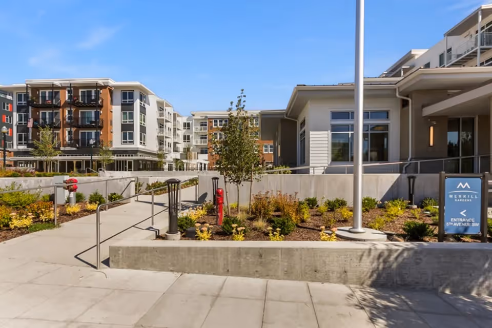 Exterior view of Merrill Gardens at Burien senior living facility showing a landscaped entrance area with plants, a concrete walkway with handrails, and multi-story residential buildings in the background under a clear blue sky.