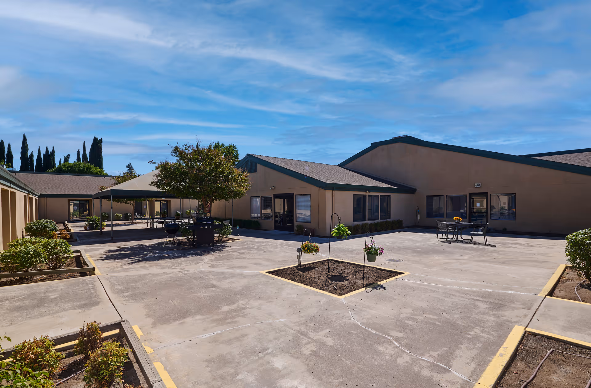 Sunny courtyard with concrete patio, planter beds, picnic tables and single-story beige buildings under a blue sky.
