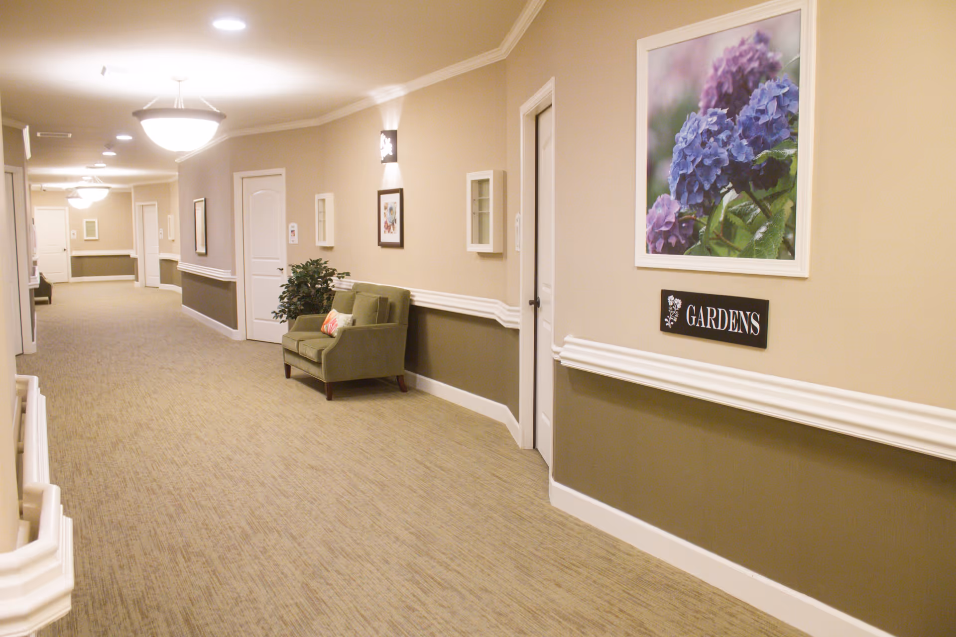 A well-lit hallway in a senior living facility with beige walls and carpeted floor. The hallway features white doors, framed floral artwork, a green armchair with a decorative pillow, and a potted plant. A sign on the wall reads 'GARDENS'.