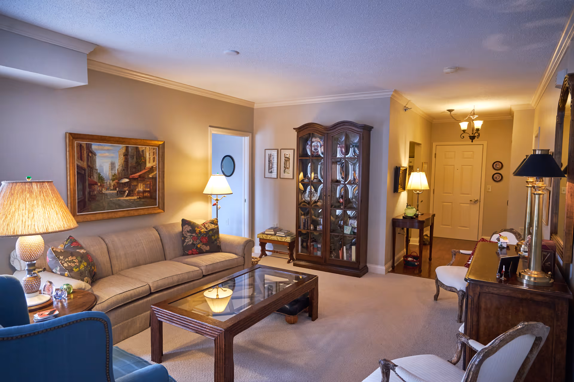 Cozy furnished living room with a sofa, glass-top coffee table, display cabinet, lamps and framed artwork.