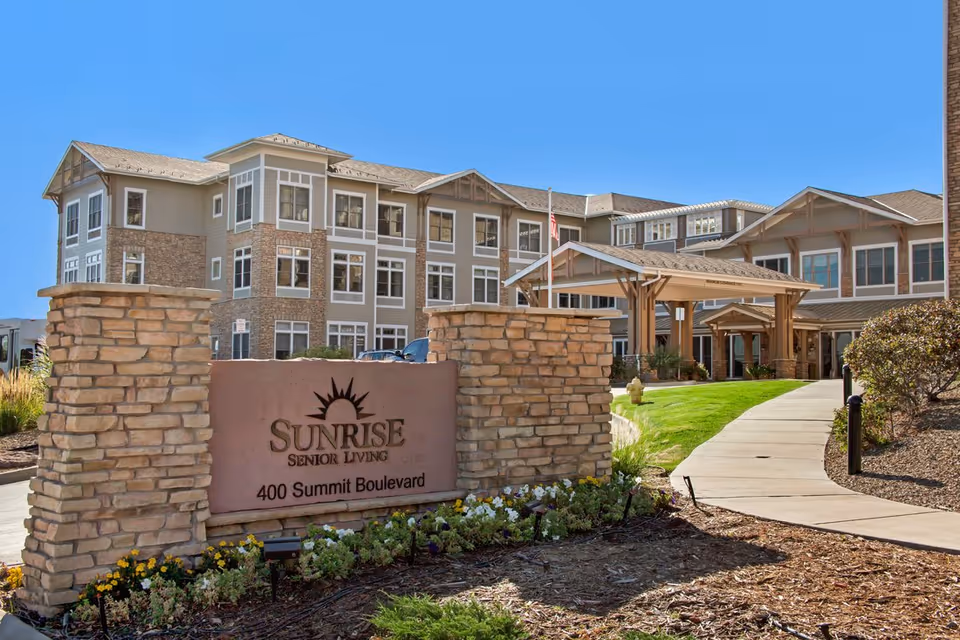 Entrance of the Sunrise Senior Living building with a stone sign reading 'Sunrise Senior Living' and a landscaped pathway leading to the covered entry.