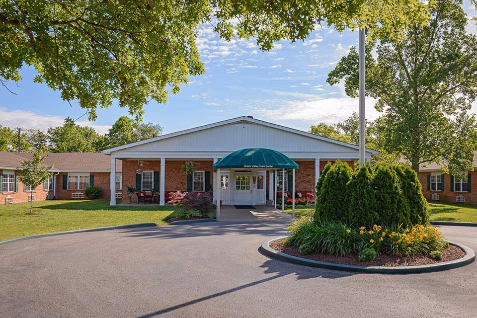 Front entrance of a single-story brick care facility with a green awning that reads "Green Valley Care Center", a circular driveway, and landscaped greenery.