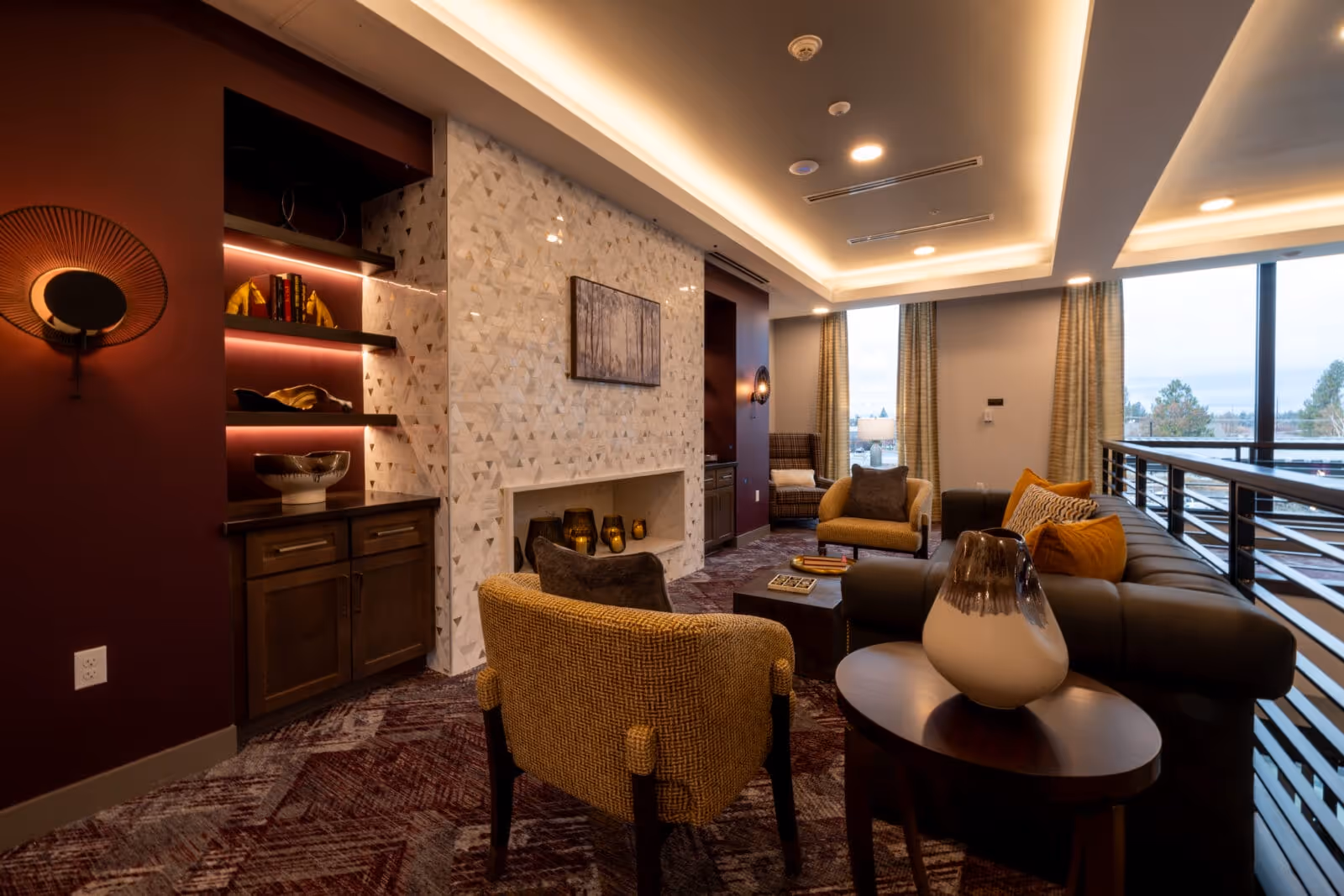 A cozy and modern living room area in a senior living facility featuring comfortable armchairs and a sofa with decorative pillows, a small round table with a ceramic vase, a built-in fireplace with candles, shelves with decorative items, large windows with curtains allowing natural light, and warm ambient ceiling lighting.