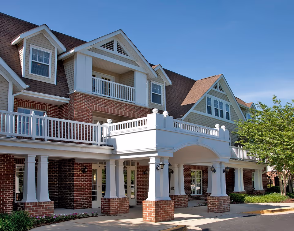 Exterior view of a senior living facility named The Willows, featuring a brick and beige siding building with white railings and columns, a covered entrance, and a clear blue sky.