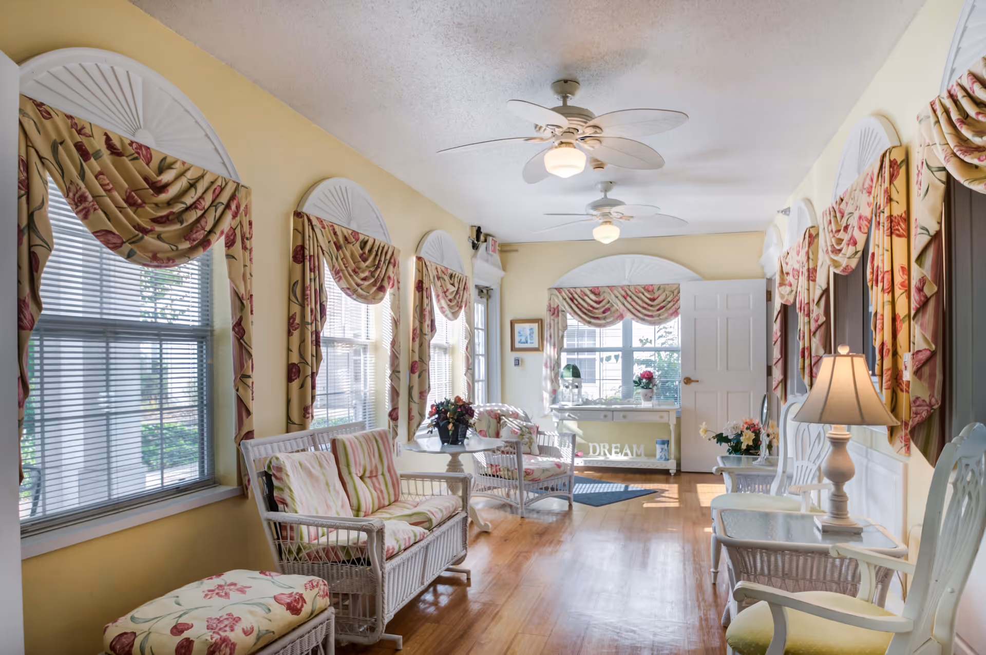 A bright and cozy sitting area with yellow walls and multiple windows adorned with floral curtains. The room features wicker furniture with floral cushions, a small table with a flower arrangement, a lamp on a side table, and ceiling fans. A white door and a small table with decorative items including a sign that says 'DREAM' are visible at the far end.