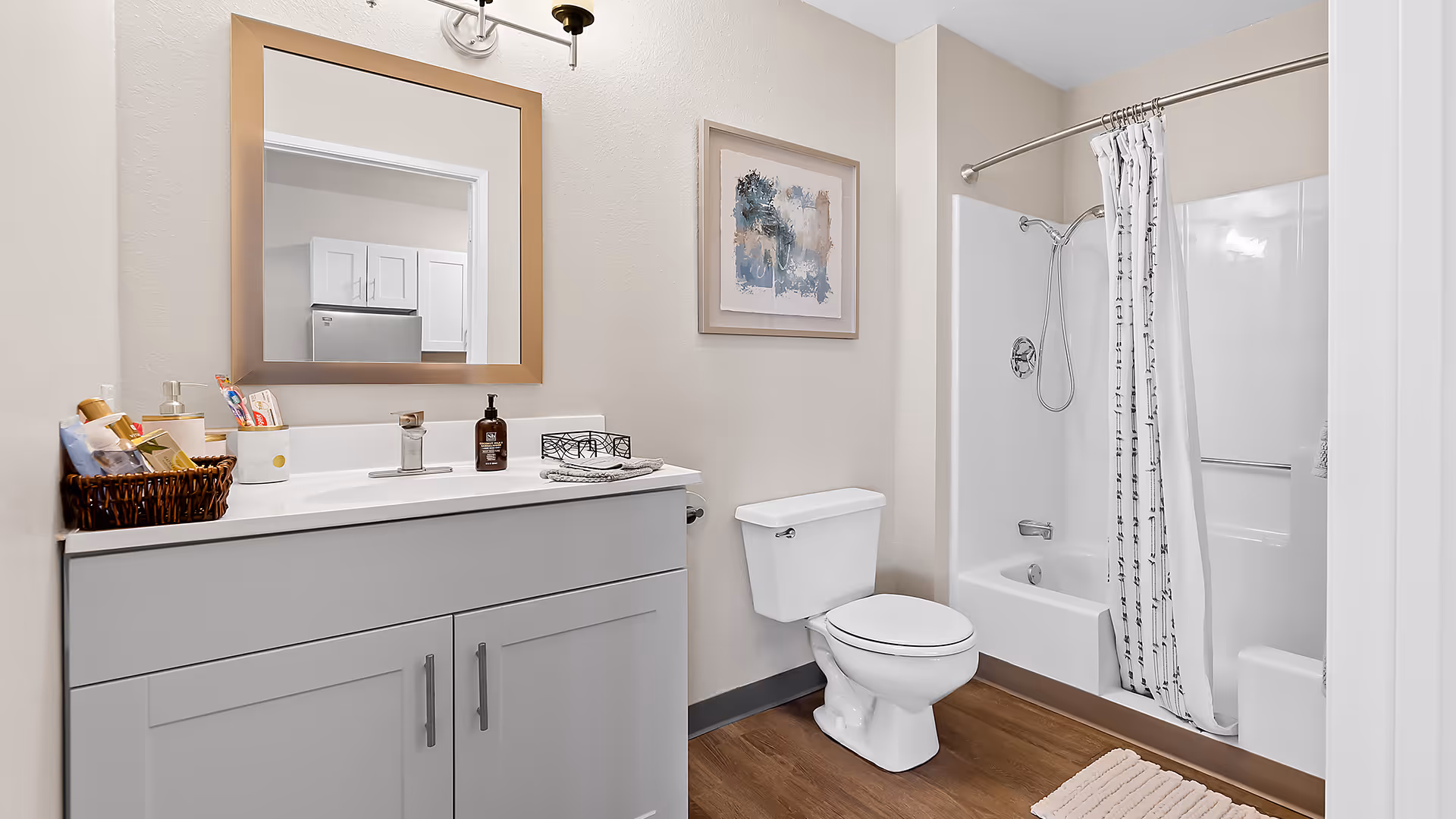 A clean and modern bathroom featuring a white vanity with a large mirror above it, a basket with toiletries on the counter, a toilet, and a bathtub with a showerhead and a white shower curtain with a black pattern. The floor is wood-style, and there is a framed abstract painting on the wall.