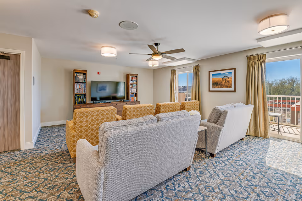 Bright common living room with upholstered sofas and armchairs facing a TV and bookshelves, a ceiling fan overhead, and a sliding glass door to a balcony.
